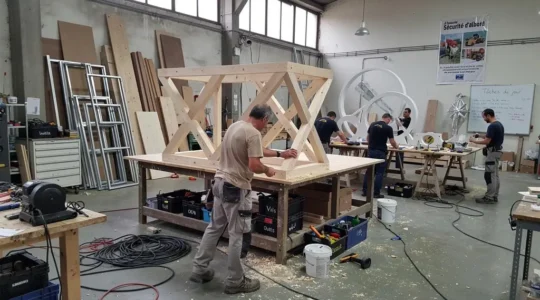 Atelier de fabrication de décors événementiels où un menuisier assemble une structure en bois sur un établi, tandis que d'autres artisans travaillent en arrière-plan sur différentes créations scénographiques.